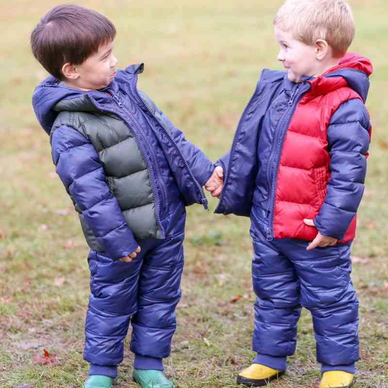 Two children wearing navy Packable Snow Pants, one paired with a blue Road Coat and the other with a red Road Coat, holding hands outdoors.